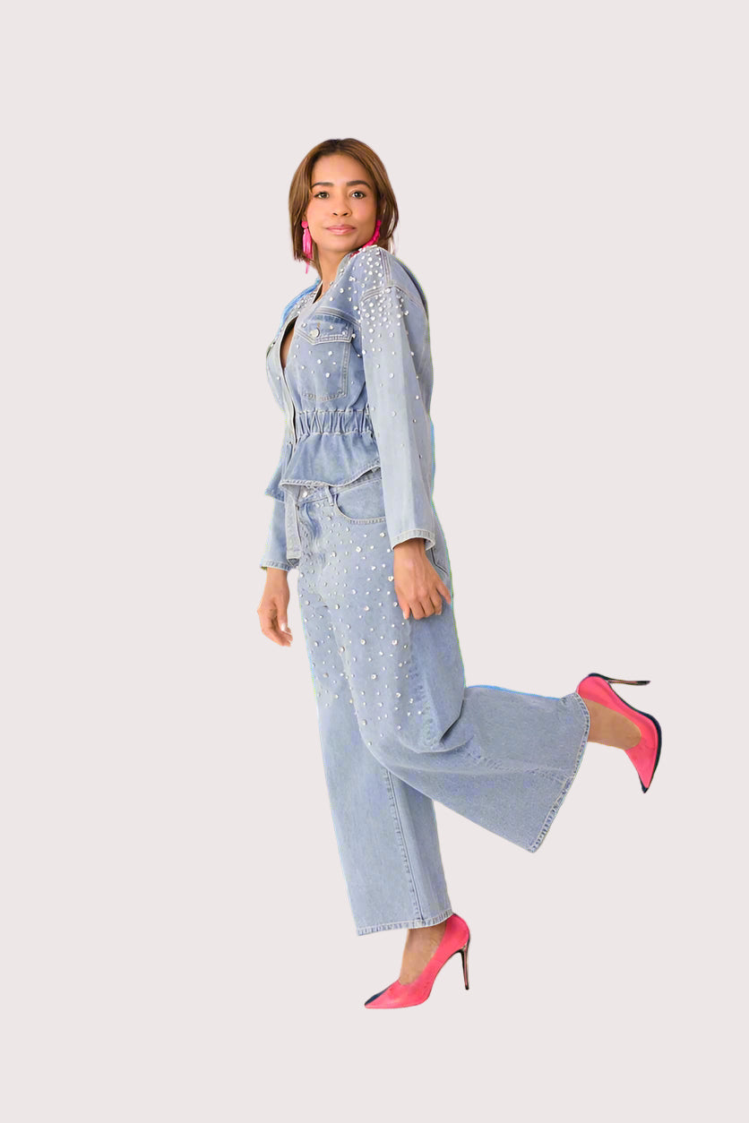 Pearl Cascade peplum denim jacket style 82325 in denim shown in a playful front walking pose with leg lift highlighting the jacket’s movement, pearl embellishments, and modern denim styling by For Her NYC