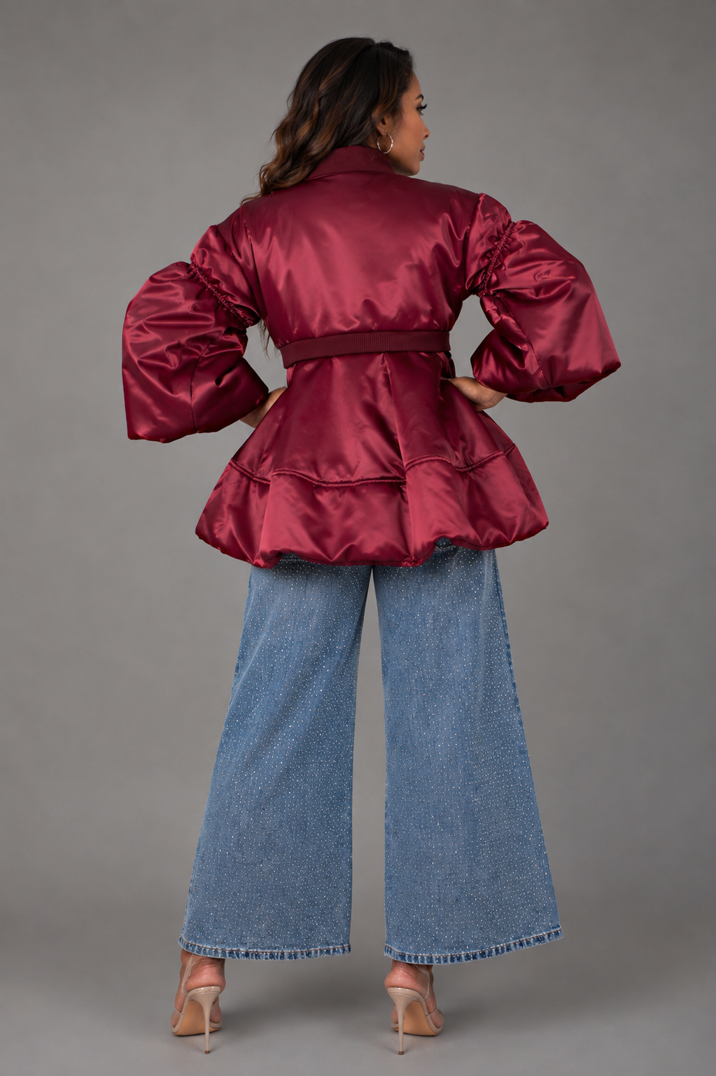 A woman models from the back a burgundy bomber jacket with dramatic puffed balloon sleeves and a matching waist belt, paired with wide-leg blue jeans and clear high heels, against a solid gray studio background.