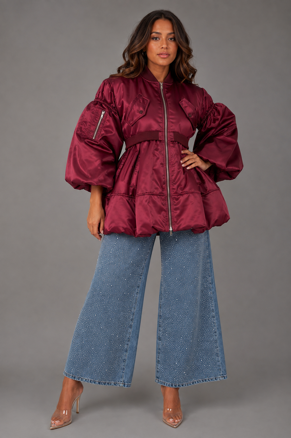A woman models a burgundy bomber jacket with dramatic puffed balloon sleeves and a matching waist belt, paired with wide-leg blue jeans and clear high heels, against a solid gray studio background.