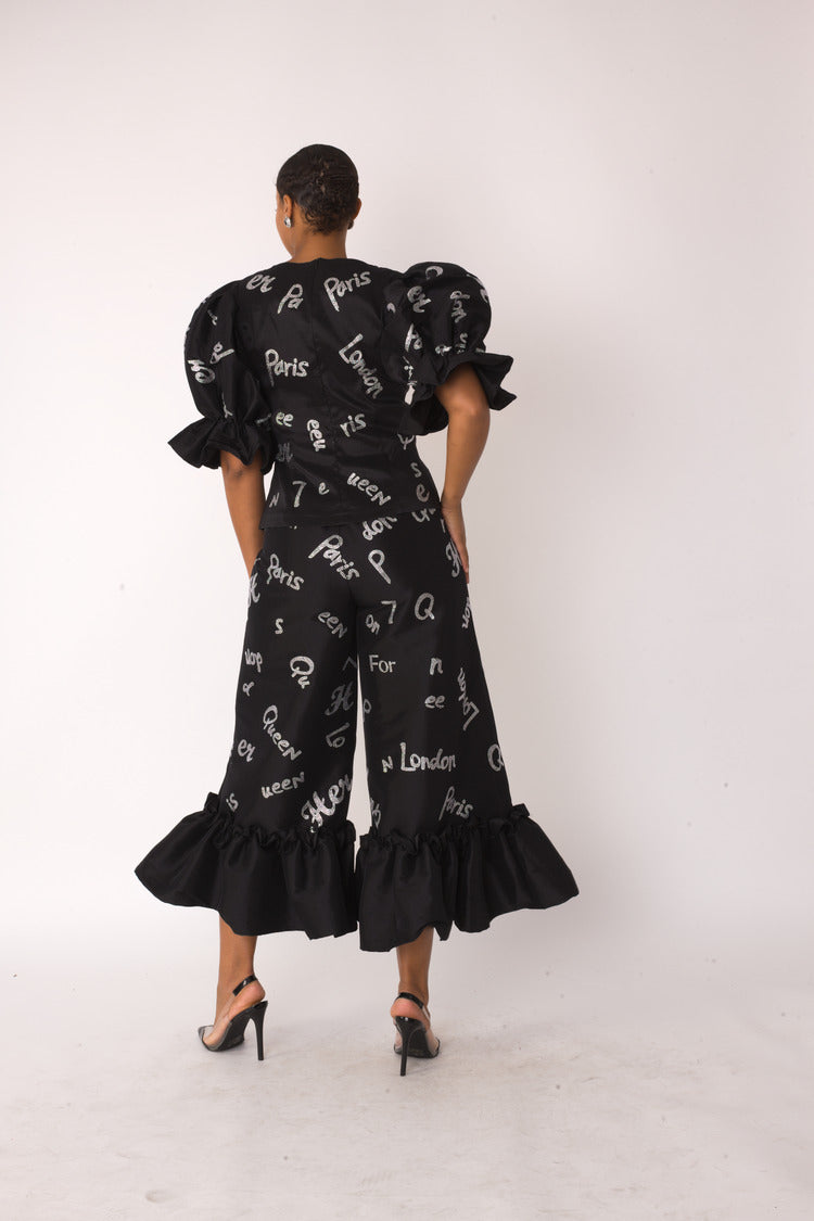 City Lights ruffle set style 82383 in black and silver shown from the back in a standing pose highlighting the allover city text print, puff sleeves, and ruffle hem detail by For Her NYC