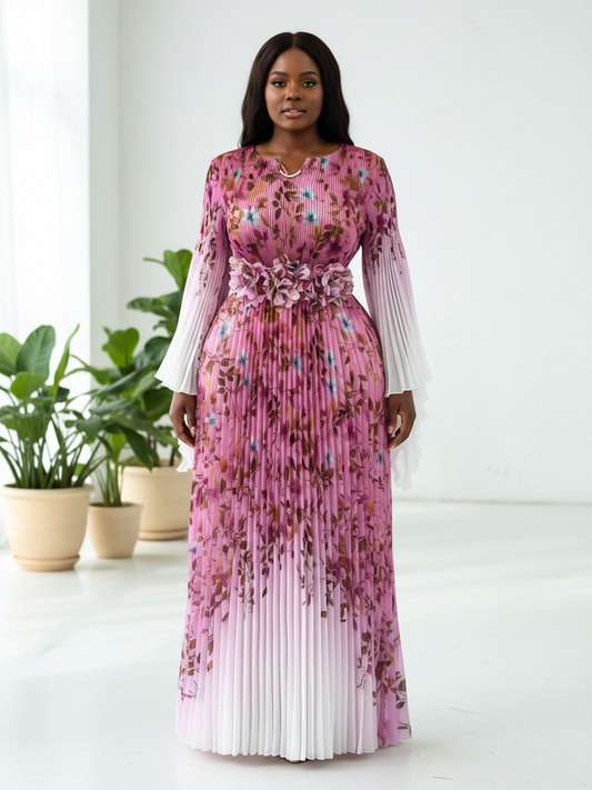 Pink multi floral pleated maxi dress with statement waist detail and sheer long sleeves modeled in a front-facing pose against a bright studio background For Her NYC