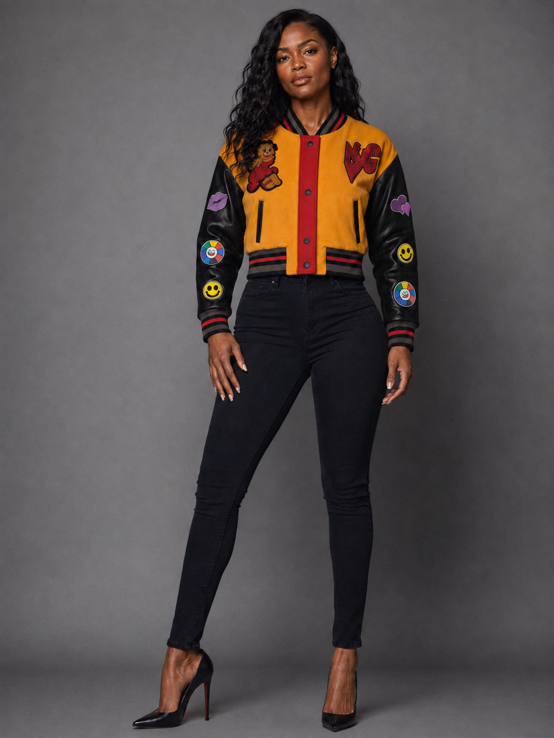 A woman modeling a Orange/Black varsity-style bomber jacket with black leather sleeves, a mustard yellow body, red trim, and various colorful patches, paired with black pants and black high heels against a gray studio background.