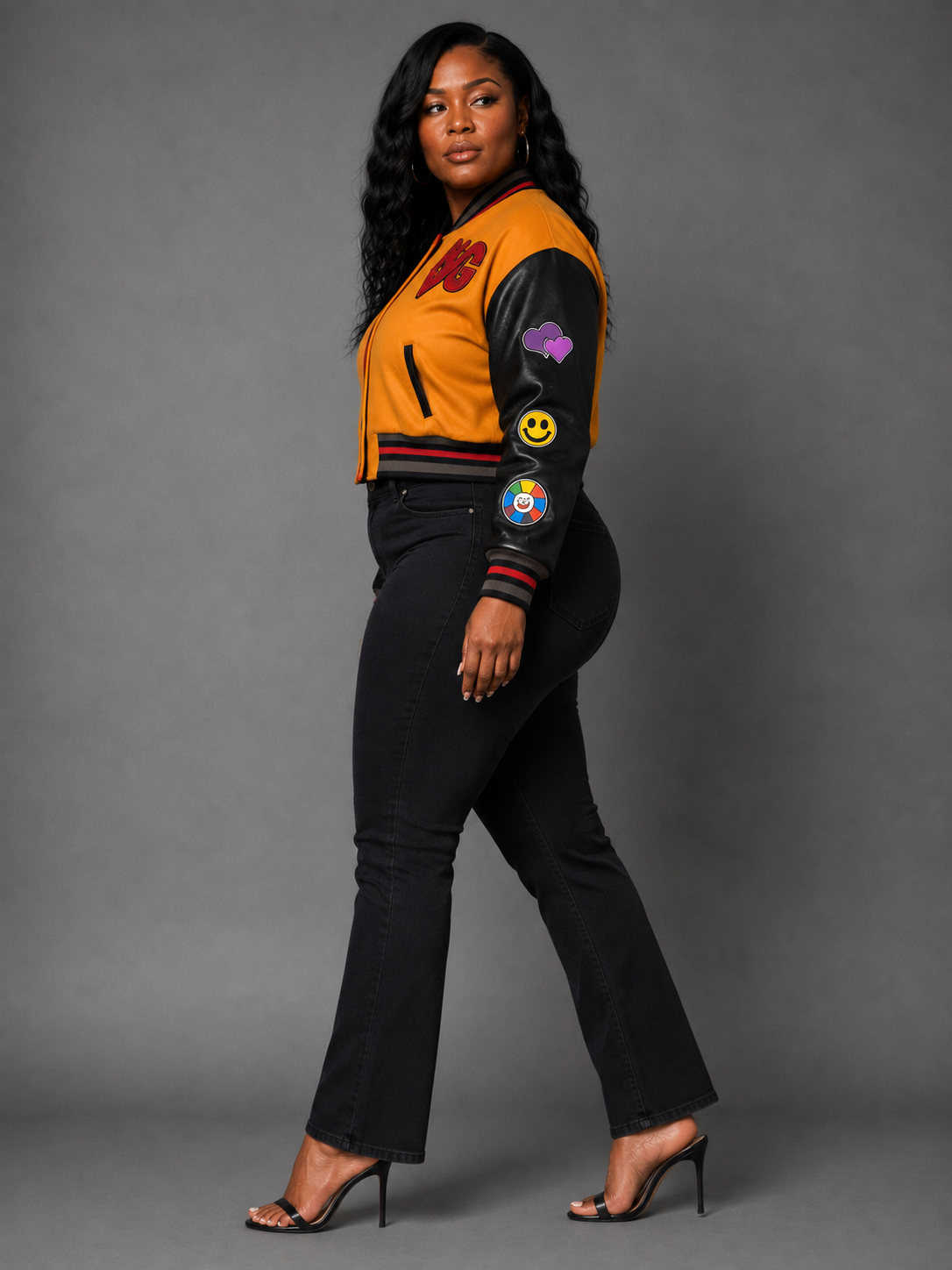 A woman modeling a Orange/Black varsity-style bomber jacket with black leather sleeves, a mustard yellow body, red trim, and various colorful patches, paired with black pants and black high heels against a gray studio background.
