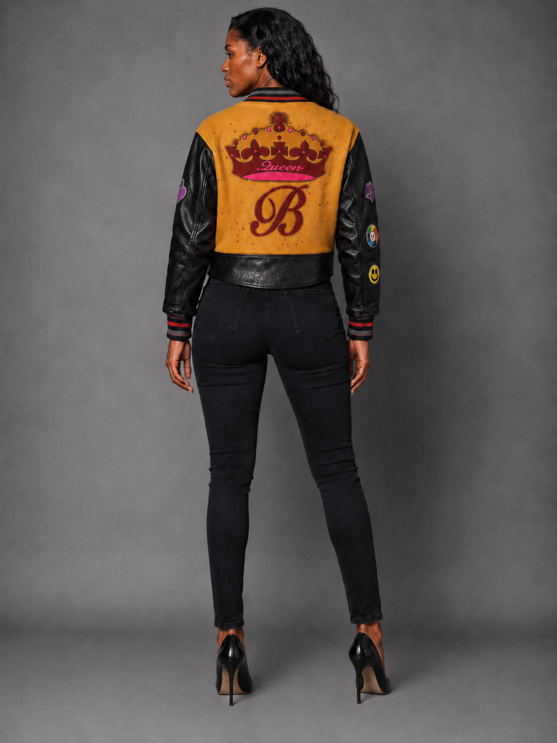 A woman modeling a Orange/Black varsity-style bomber jacket with black leather sleeves, a mustard yellow body, red trim, and various colorful patches, paired with black pants and black high heels against a gray studio background.