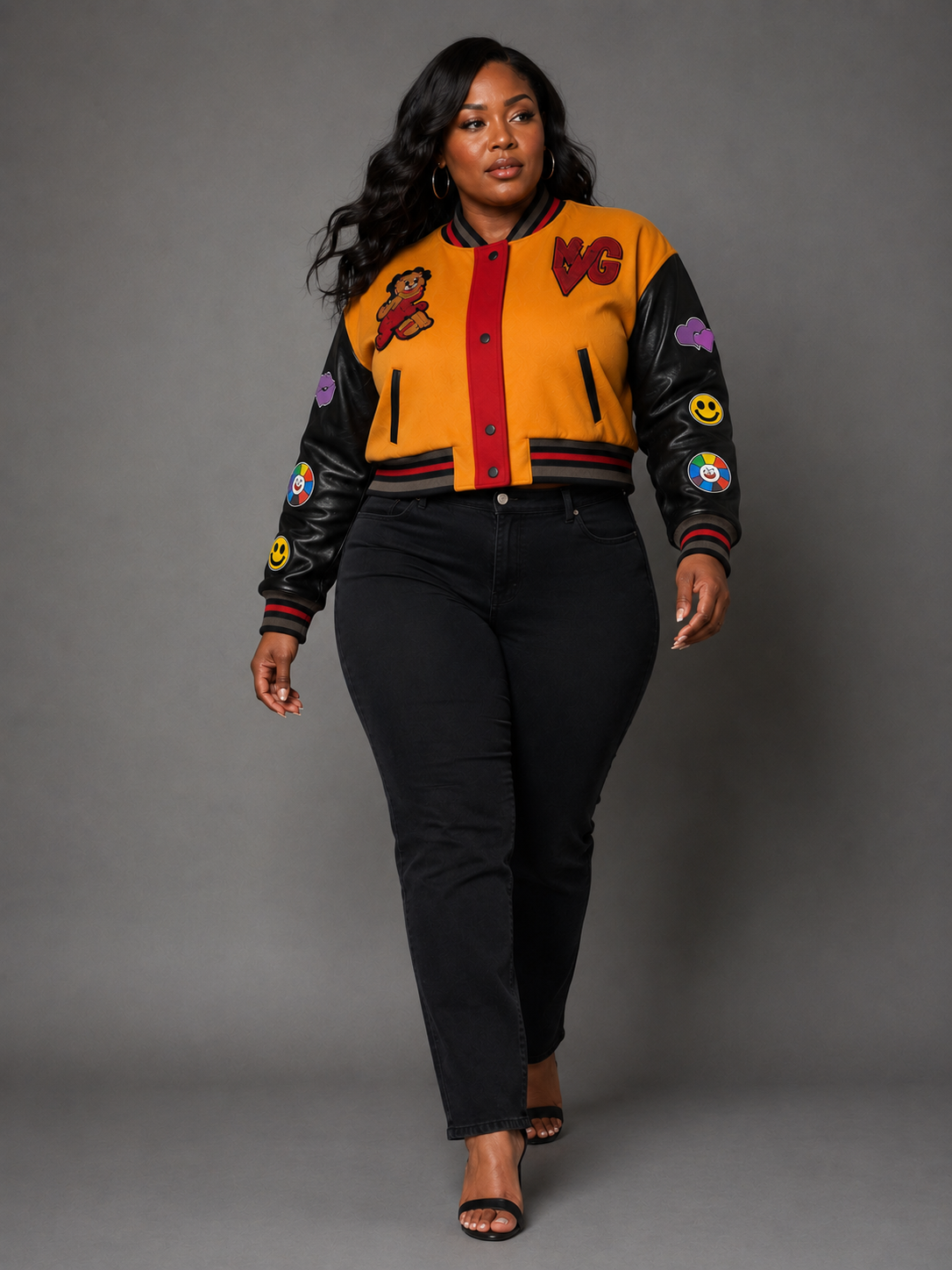 A woman modeling a Orange/Black varsity-style bomber jacket with black leather sleeves, a mustard yellow body, red trim, and various colorful patches, paired with black pants and black high heels against a gray studio background.