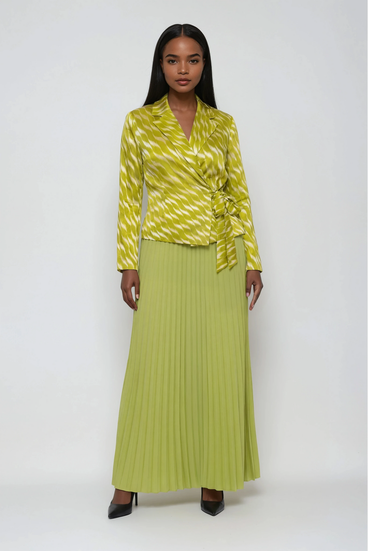 Woman wearing a yellow pleated midi skirt with a matching structured wrap-style jacket and tie waist, styled with heels — For Her NYC