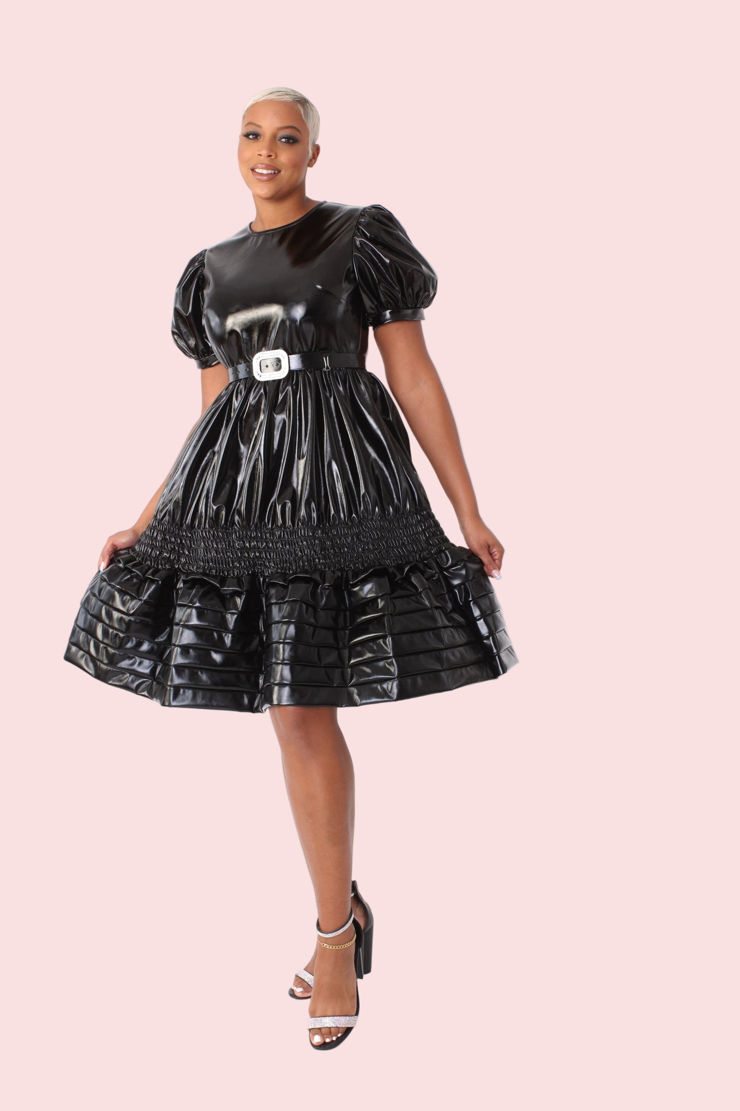 Model wearing black high gloss faux leather dress with puff sleeves and ruffle hem, front walking pose showcasing movement and glossy finish by For Her NYC