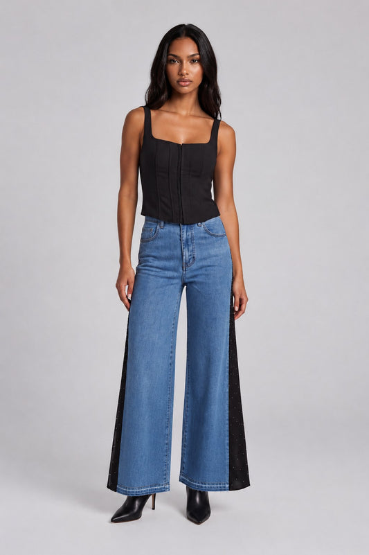 Model wearing the Crystal Drape Panel Denim Jeans featuring a medium wash denim front, black crystal-embellished contrast drape panels, a high-rise waist, and a wide-leg silhouette paired with a black camisole. For Her NYC.