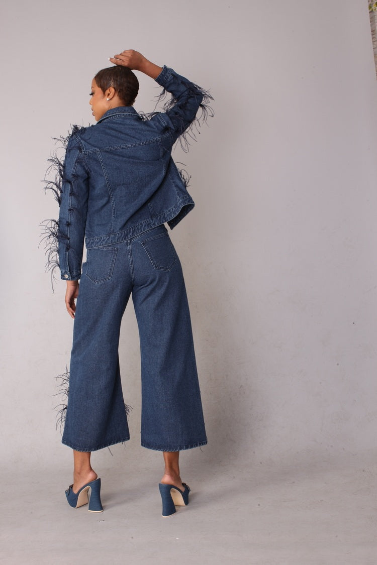 Back view of blue denim wide leg jeans with feather trim shown with hand on head pose highlighting rear fit and dramatic hem detail, For Her NYC