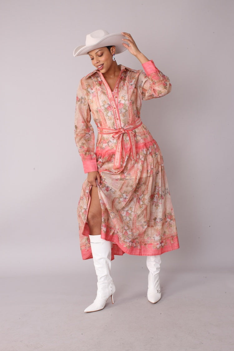 Front view of pink floral print shirt dress — collared long sleeve maxi with self tie belt and front slit, styled with white cowboy hat and white boots.