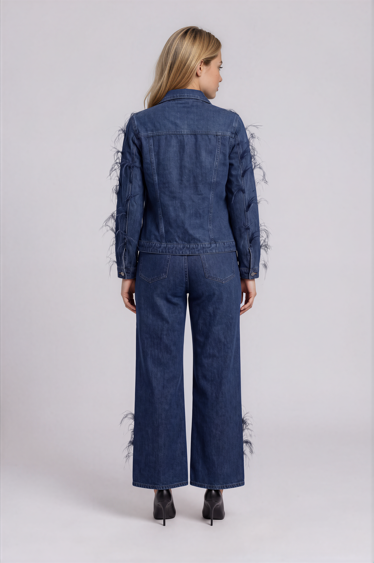 Back view of a model wearing the dark-wash For Her NYC Feather Trim Denim Jacket - 82303 with feather embellishments along the sleeves