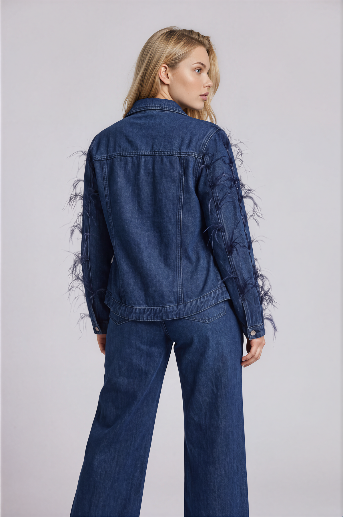 Three-quarter back view of a model wearing the dark-wash For Her NYC Feather Trim Denim Jacket - 82303 with wispy feather embellishments along the sleeves