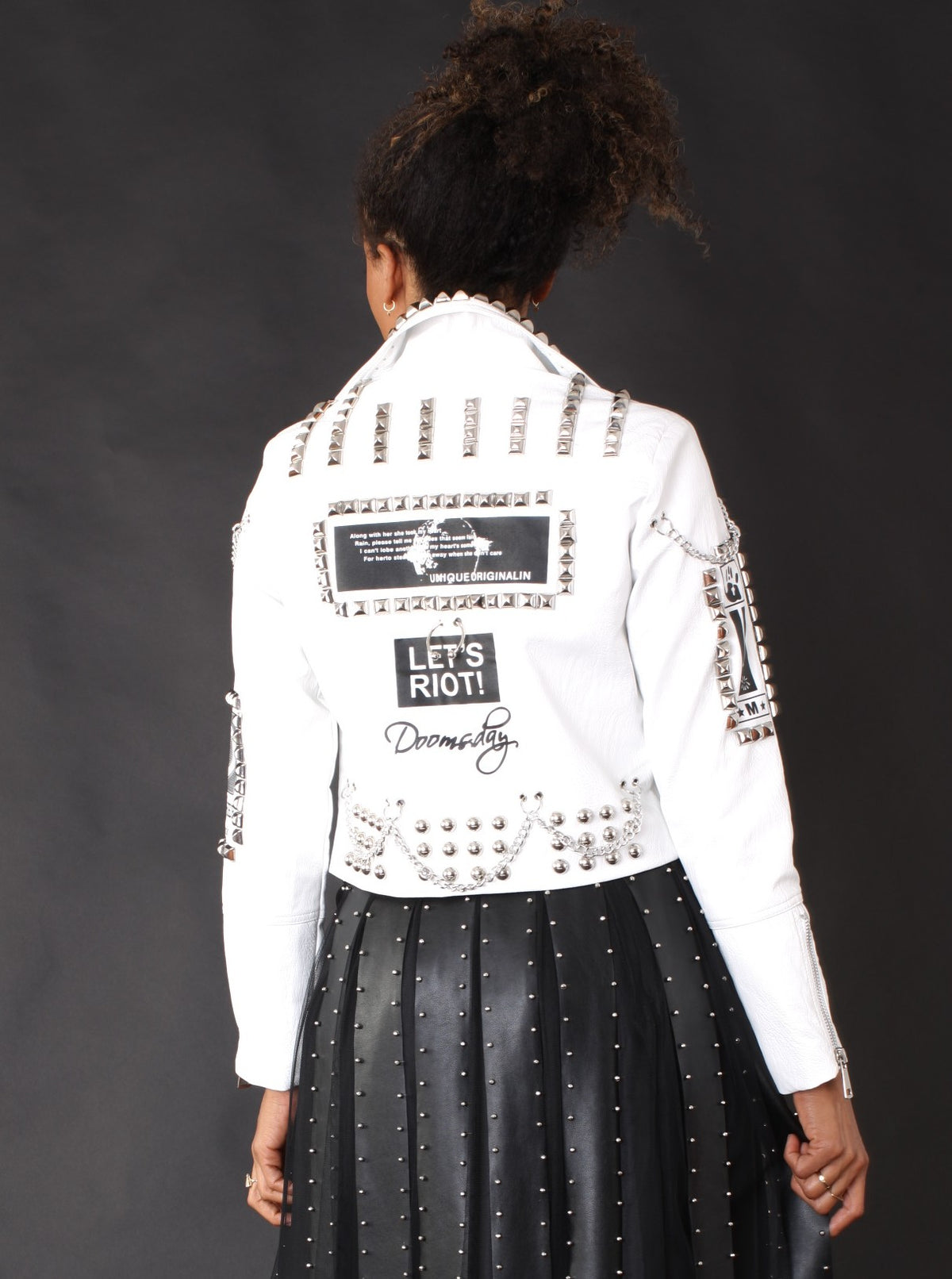 Back view of a model wearing the white For Her NYC Studded Faux Leather Moto Jacket with Graphic Prints - 81771, featuring silver studs, chains, and graphic patches.