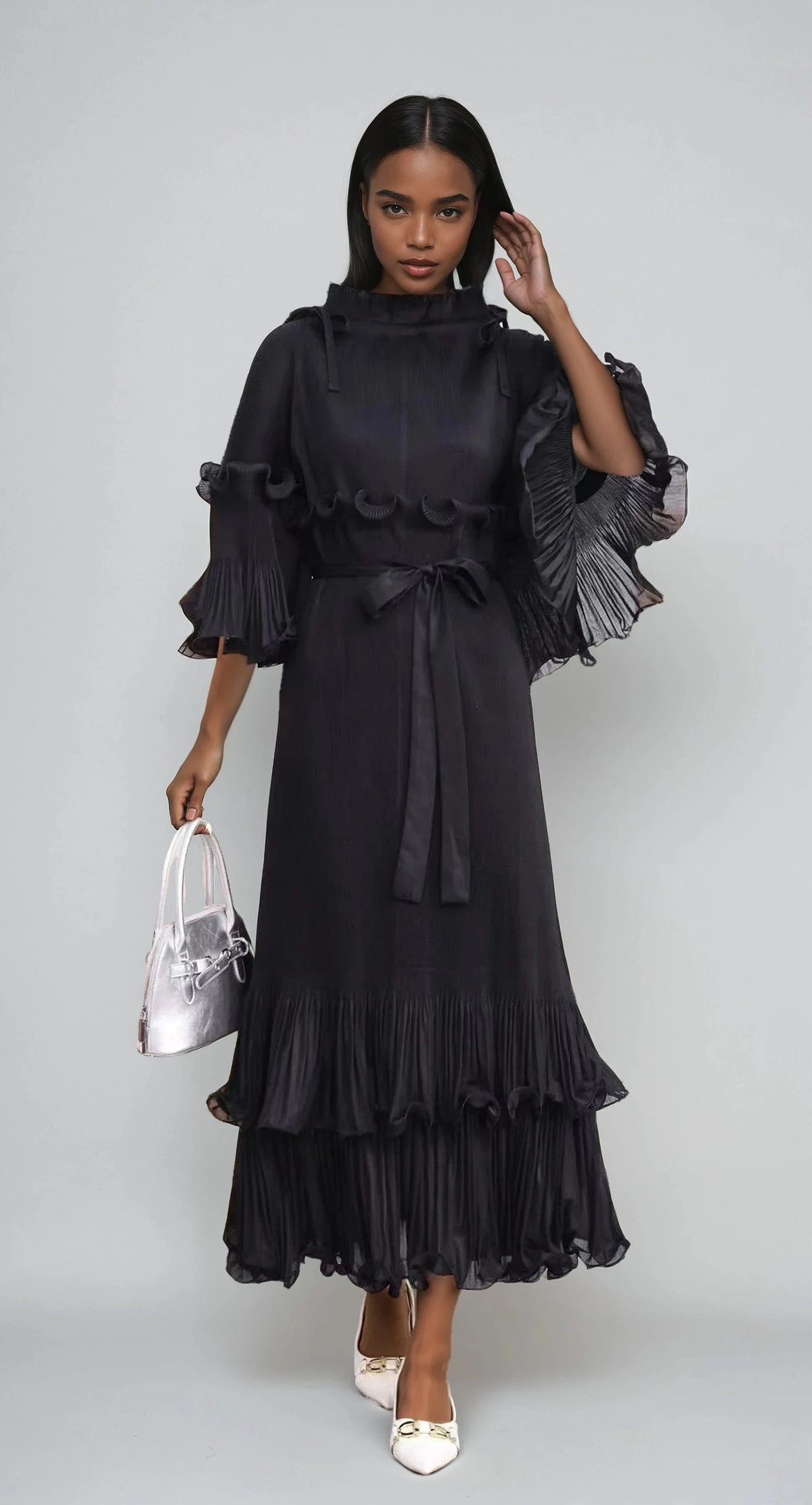 Model wearing black pleated maxi dress with ruffle sleeves, tiered hem, and belted waist by For Her NYC.
