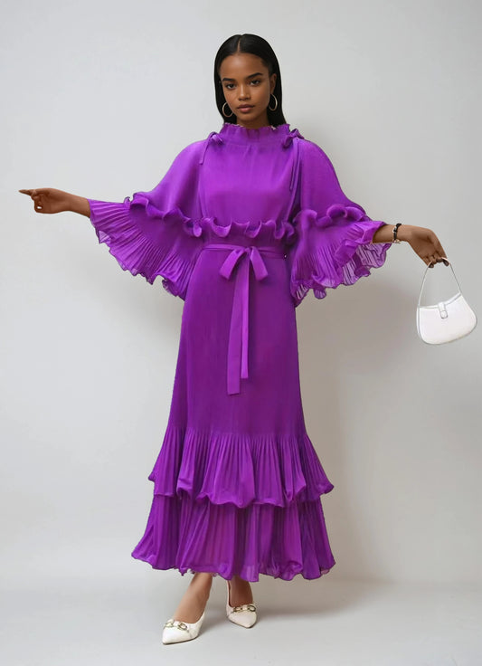 Model wearing purple pleated maxi dress with ruffle sleeves, tiered hem, and waist tie by For Her NYC.