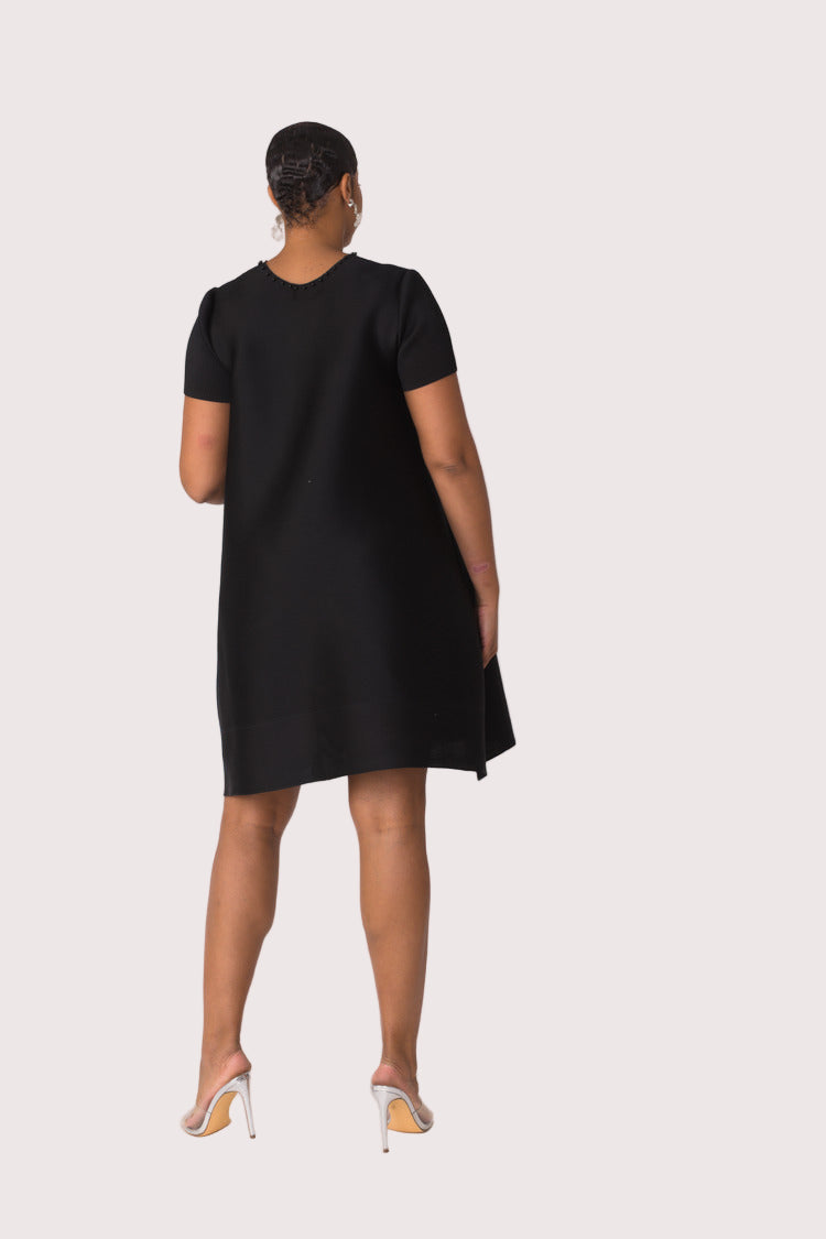 Back view of model wearing the black fringe pocket shift dress showing clean straight silhouette and knee length hem by For Her NYC