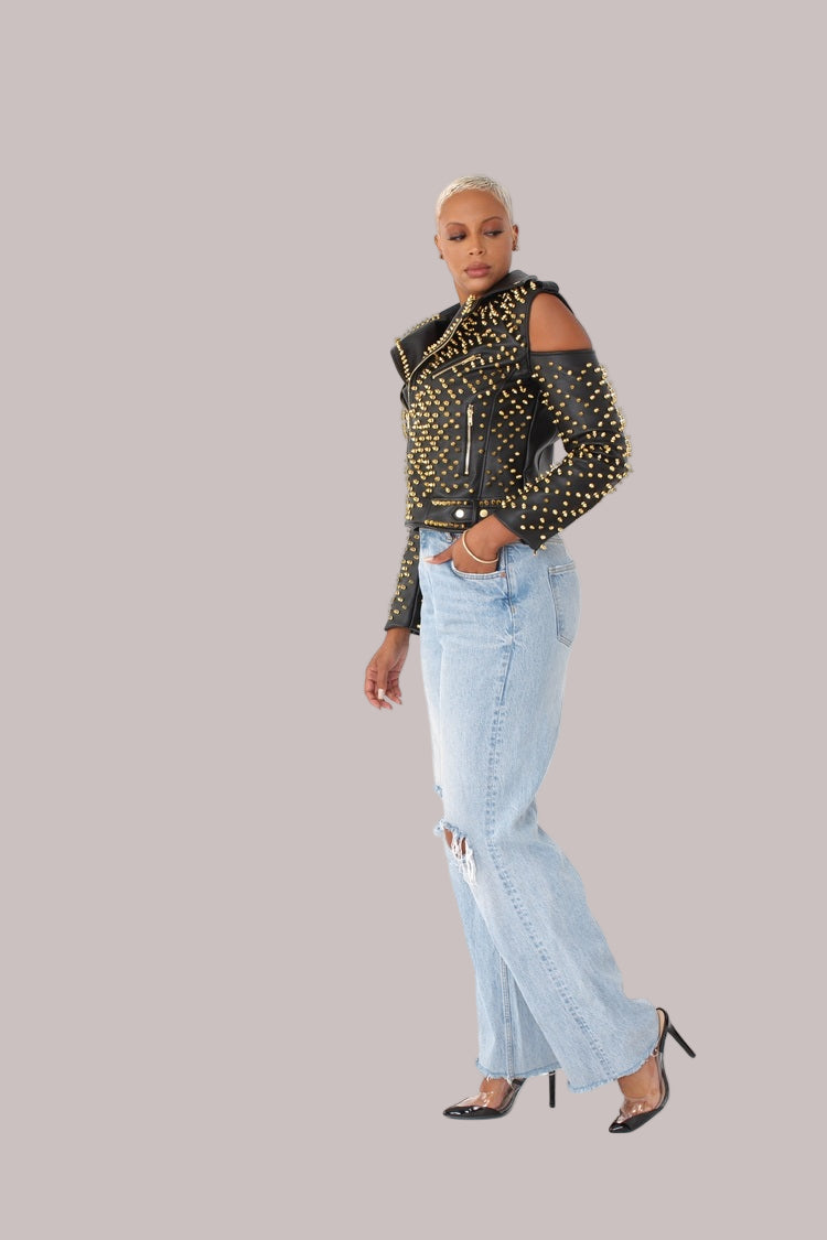 Black and gold studded pleather moto jacket with open shoulders shown in side walking view, emphasizing silhouette and stud detailing by For Her NYC